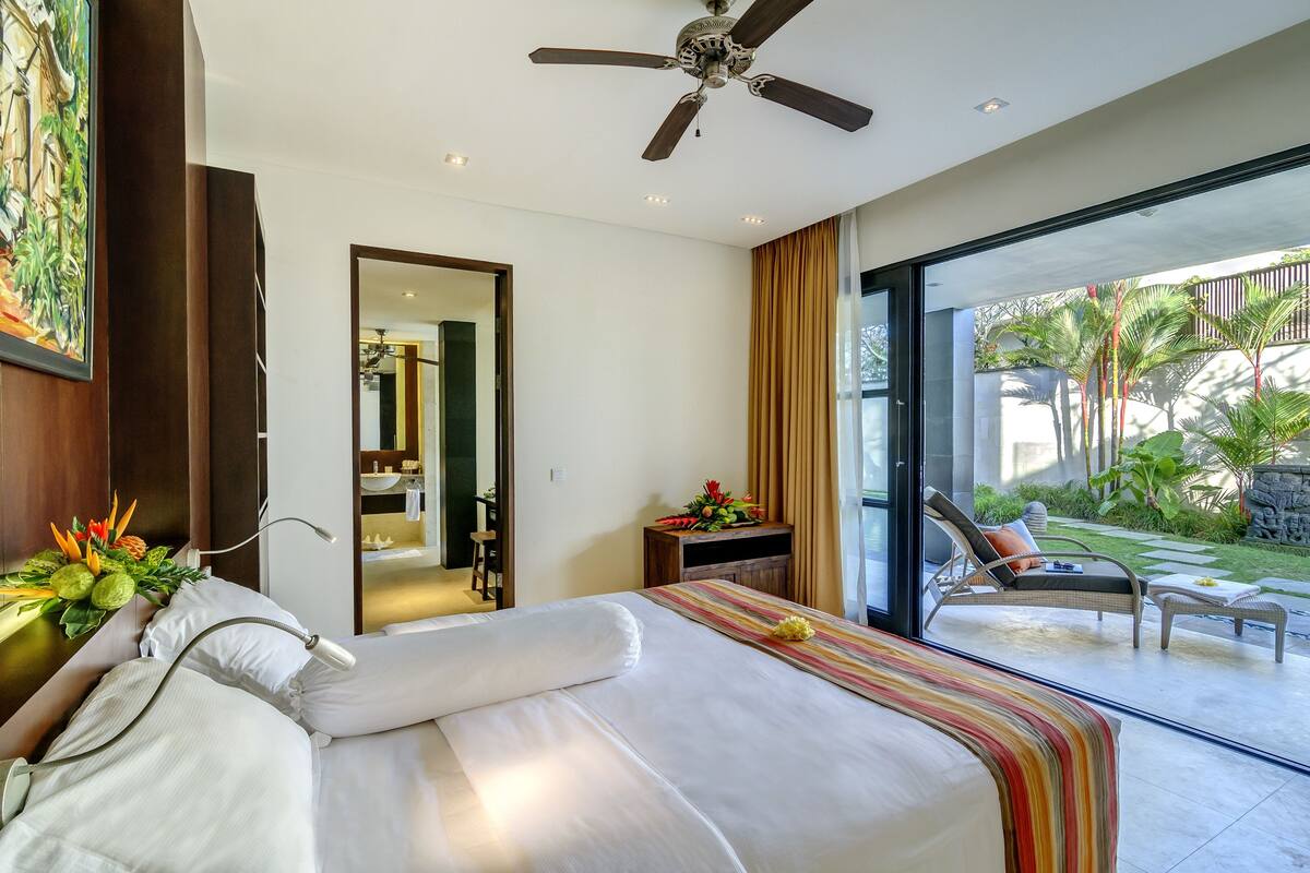 A hotel room with a bed, a ceiling fan, a large window, and a view of the outdoor garden.