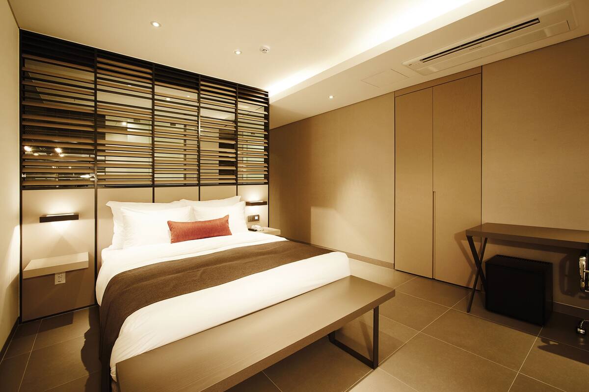 Quarto de hotel moderno com uma cama grande, luminárias, um banco, escrivaninha e guarda-roupa.
