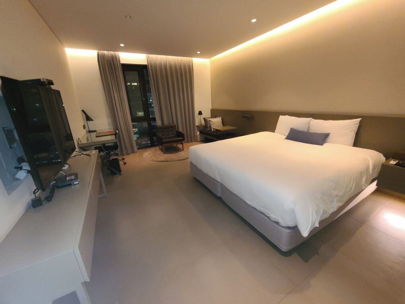 Una habitación de hotel moderna con una cama grande, un escritorio, una silla y vistas de la ciudad a través de la ventana.