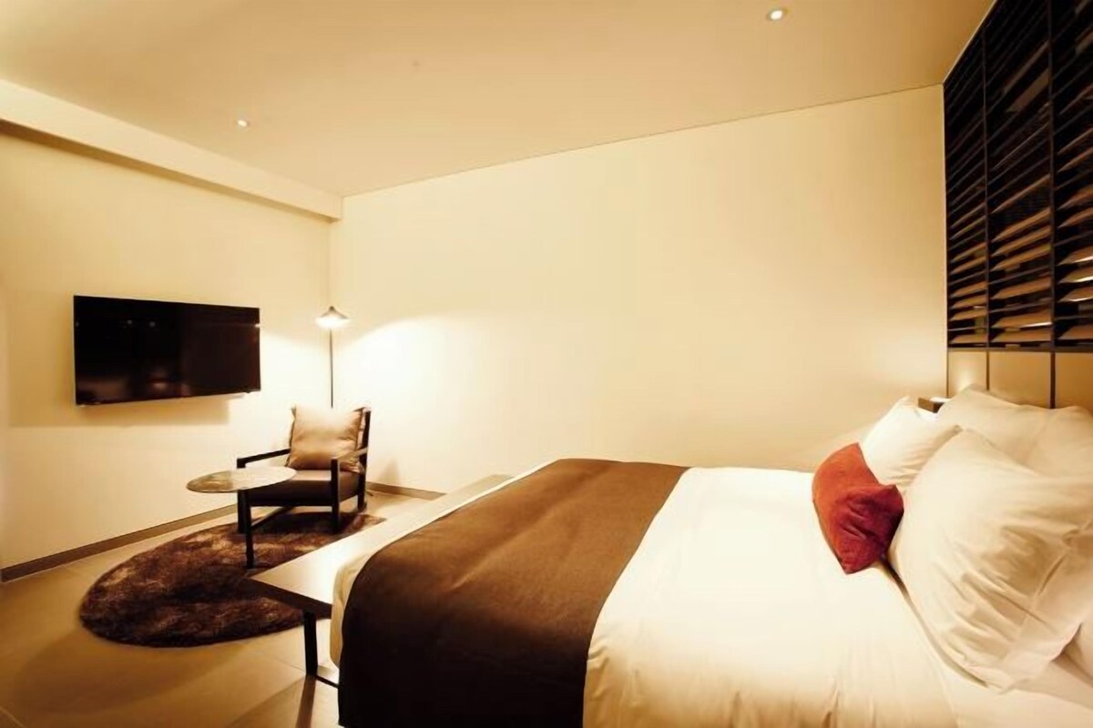 A modern hotel room with a bed, a chair, a small table, and a TV mounted on the wall.