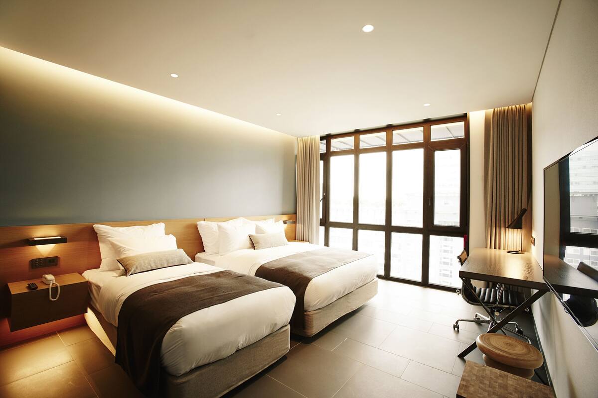 Quarto de hotel moderno com duas camas, uma escrivaninha e uma janela grande.