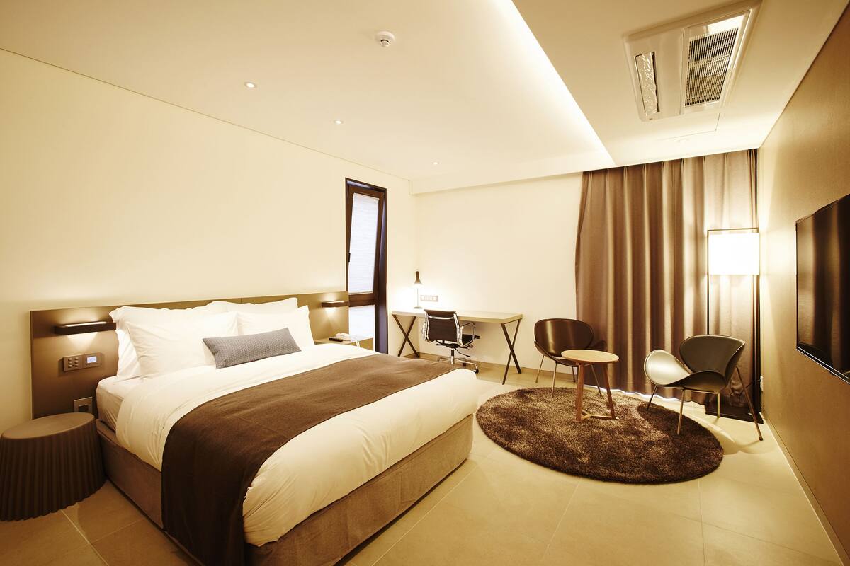 Quarto de hotel com uma cama grande, uma escrivaninha, duas cadeiras, uma televisão e um tapete redondo pequeno.
