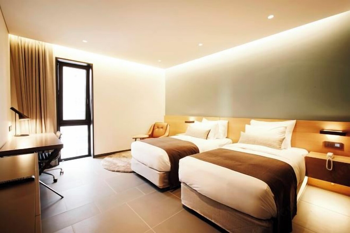 A modern hotel room with two beds, a desk, a chair, and a large window.