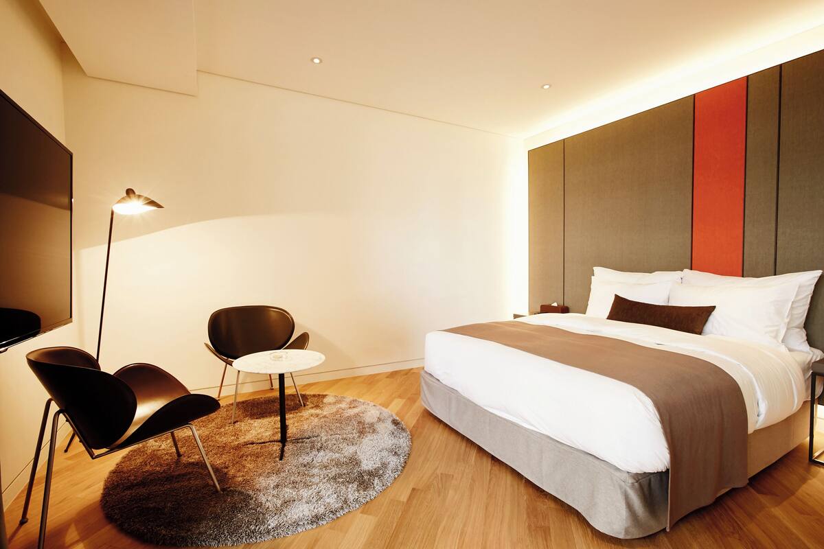 Quarto de hotel moderno com cama, duas cadeiras, uma mesa redonda pequena e um abajur.