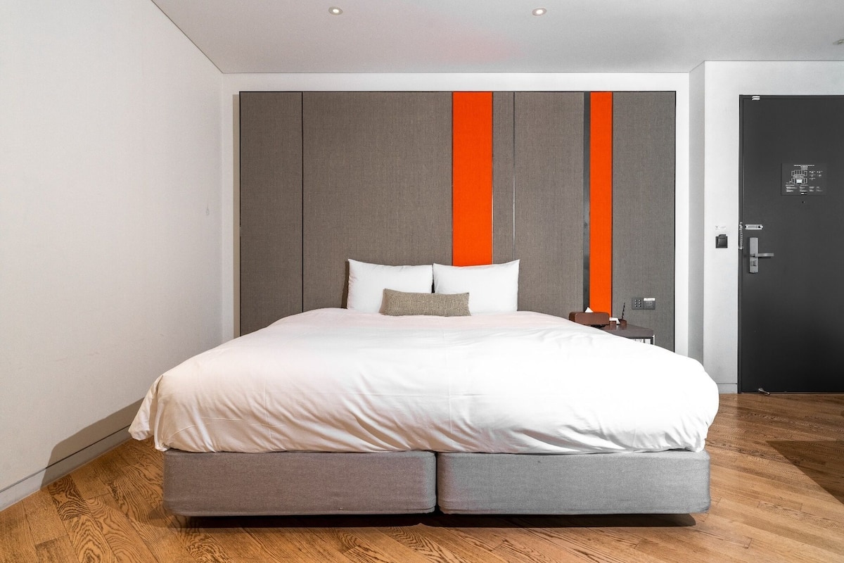 Quarto de hotel moderno com uma cama grande, painéis de parede cinza e laranja, piso de madeira e uma porta com fechadura.