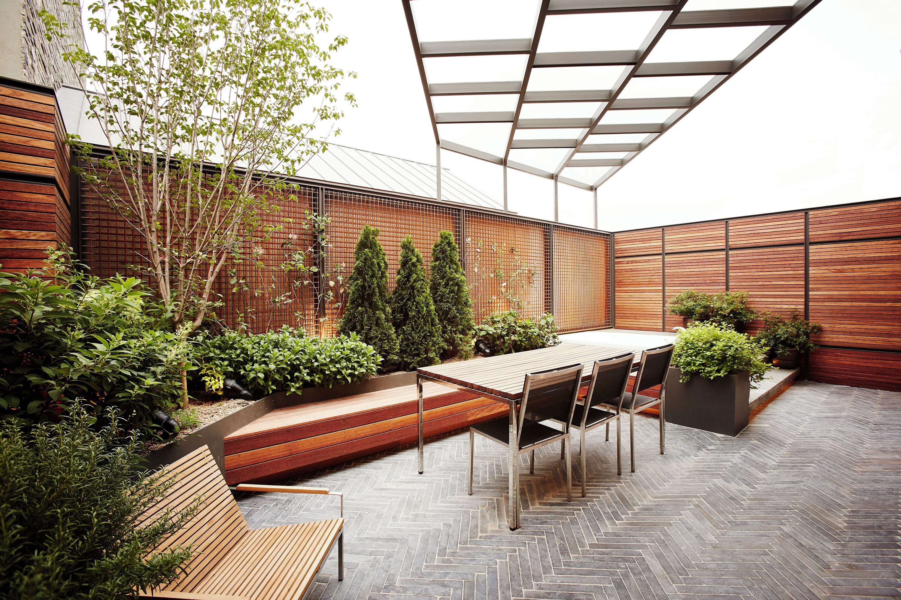 Un patio exterior moderno con una mesa y sillas de madera, rodeado de follaje y un suelo con patrón de espiga.