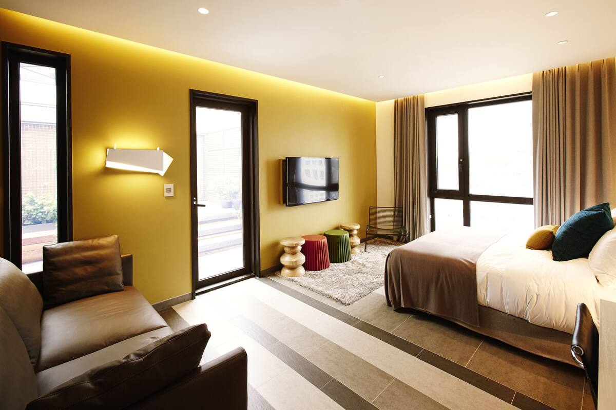 Quarto de hotel moderno com uma cama grande, um sofá, uma televisão e uma janela com cortinas.
