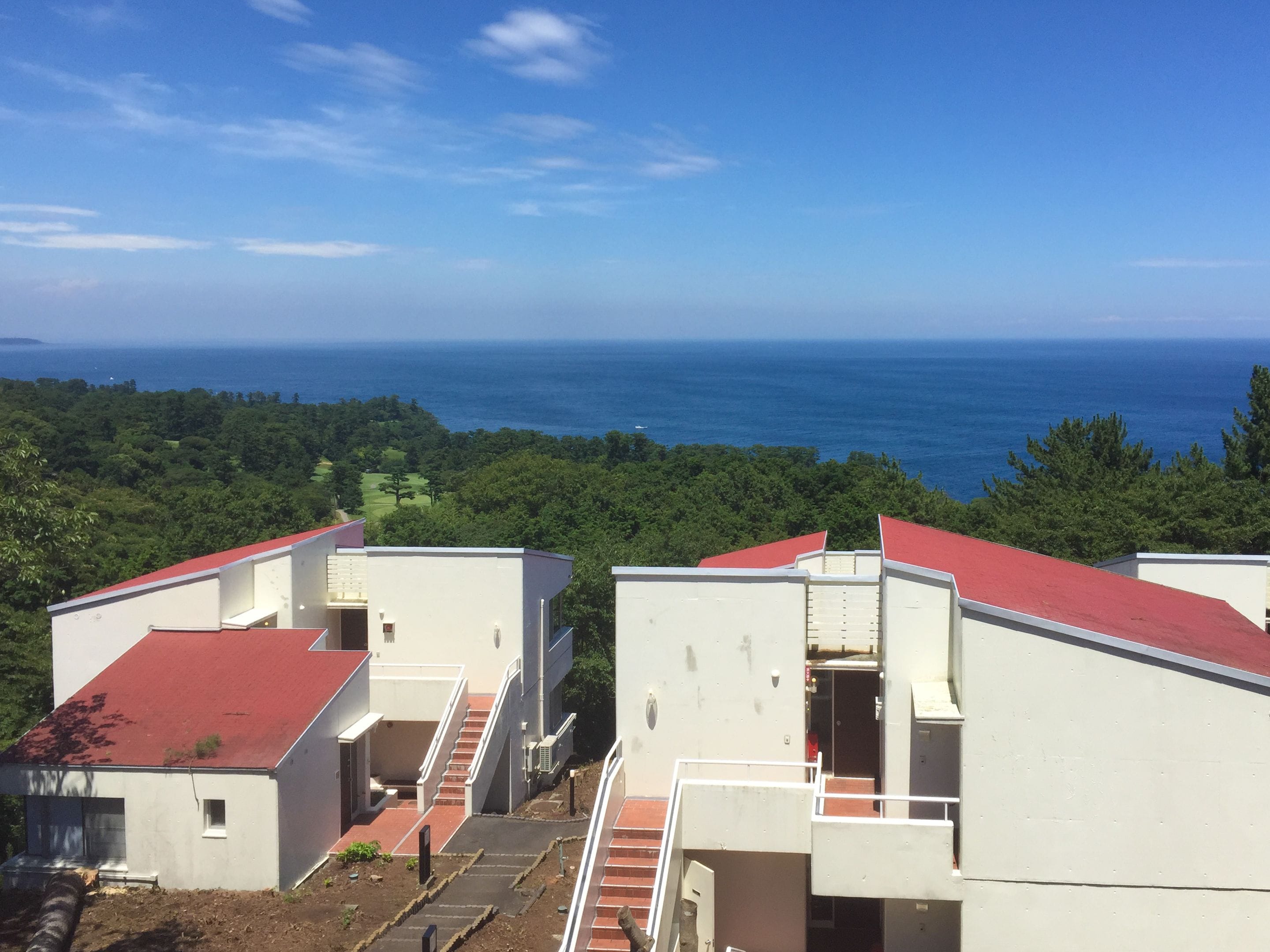 A view of a coastal building complex with a red roof, surrounded by greenery and overlooking the sea.