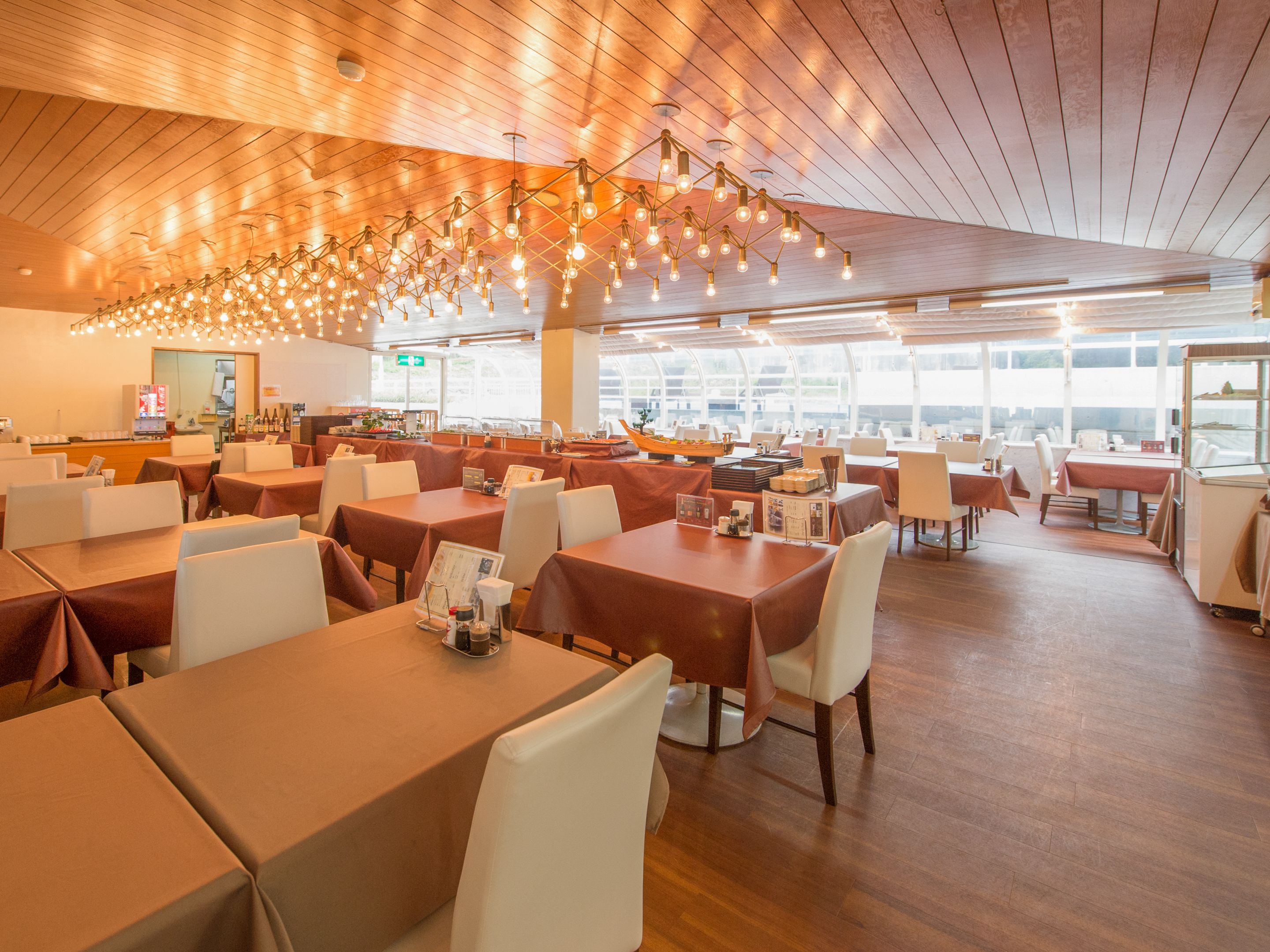 Un restaurante espacioso con numerosas mesas y sillas, techo de madera y grandes ventanales que dejan entrar la luz natural.
