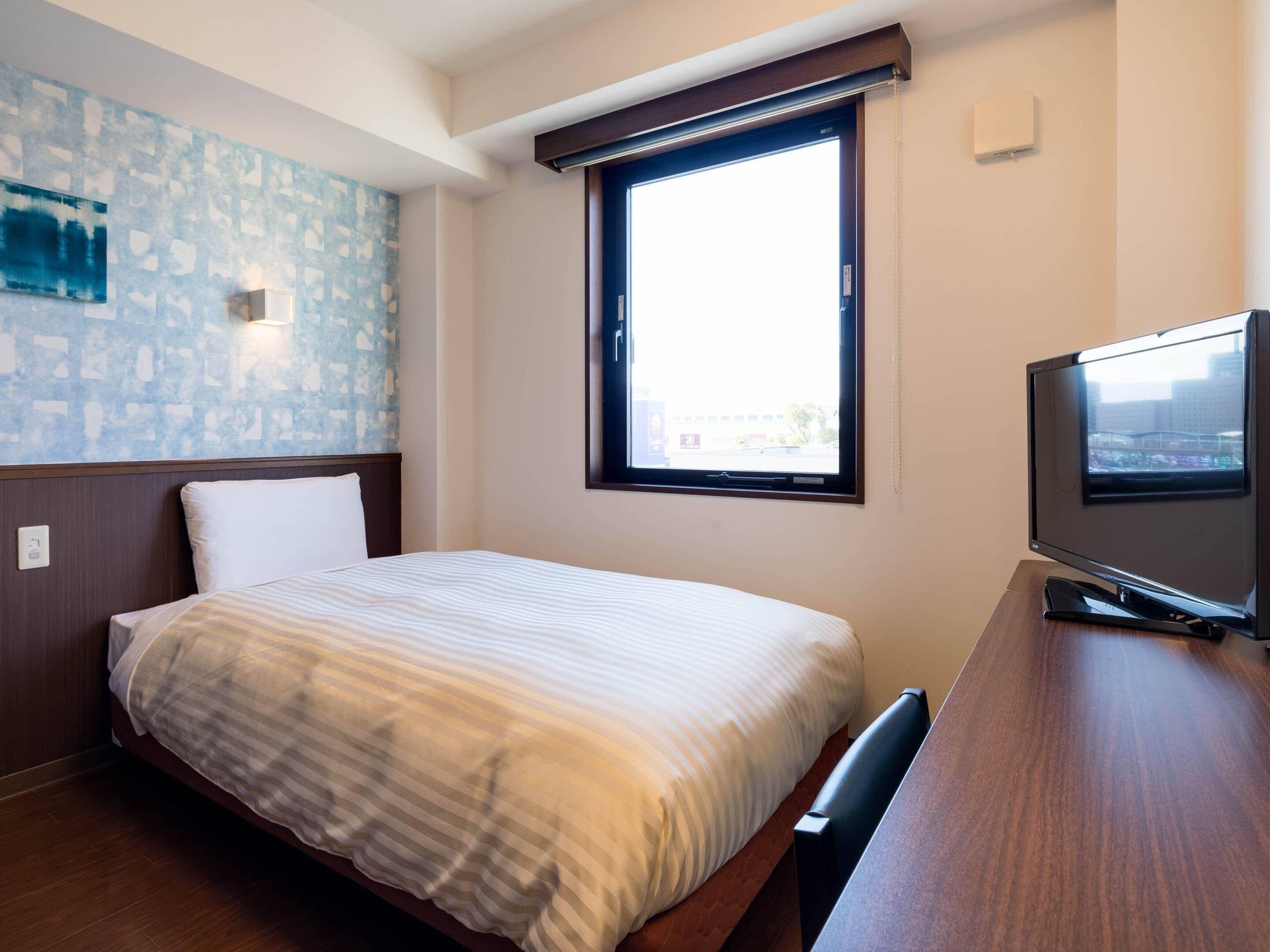 A hotel room with a bed, a television, a window with blinds, and a wall with a textured pattern.