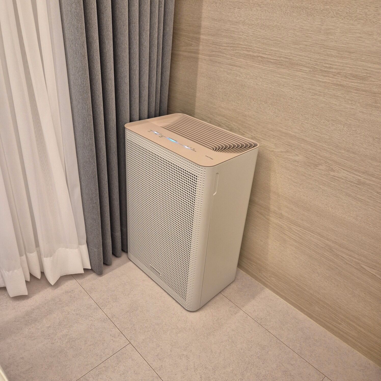 A portable air purifier in a corner of a room with a light-colored floor and a wooden wall.