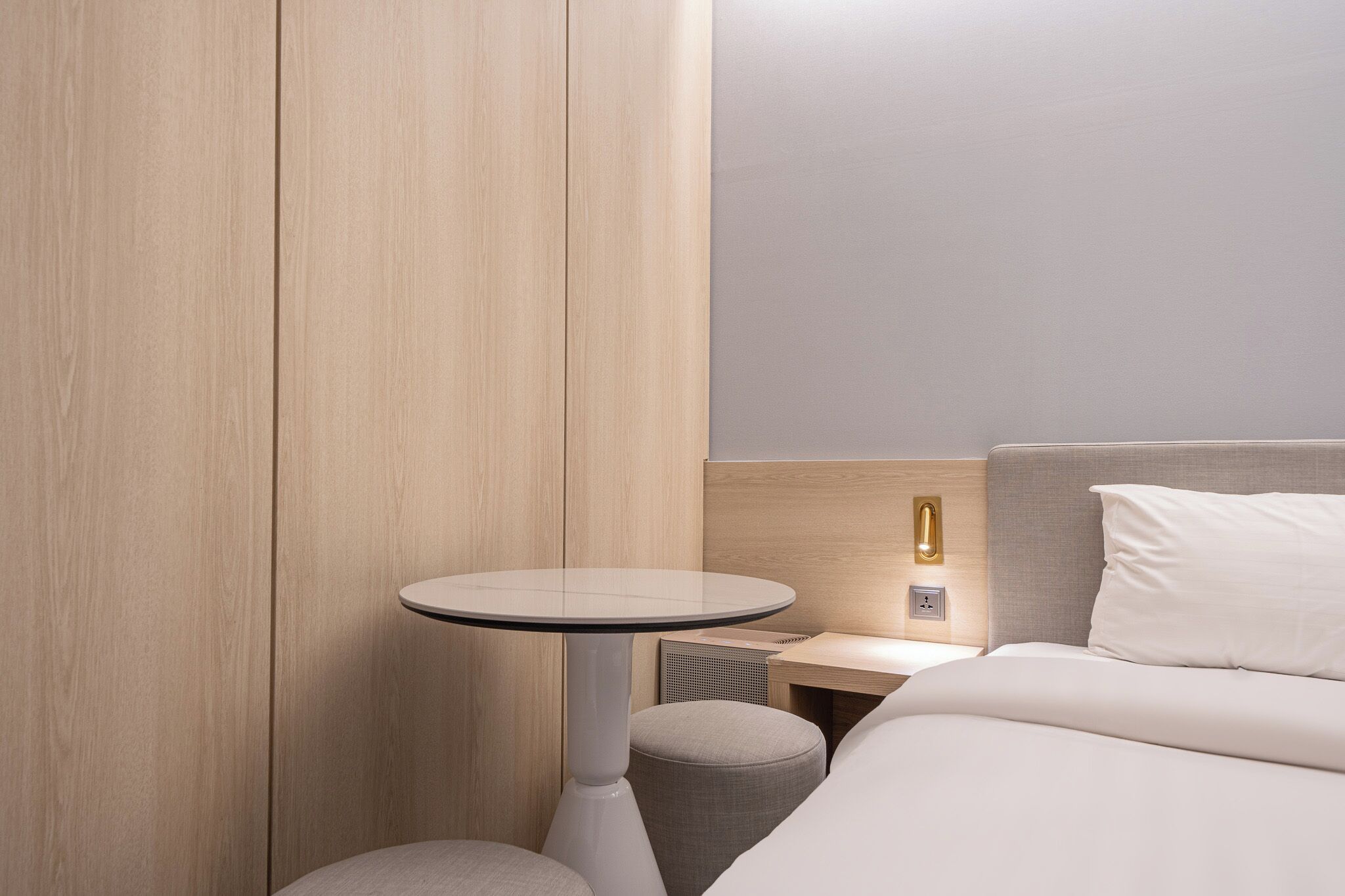 A modern hotel room with a wooden wardrobe, a round table, a bedside table with a lamp, and a bed with white bedding.