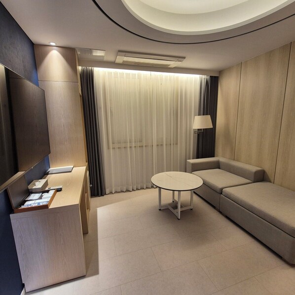 A modern hotel room with a flat-screen TV, a sofa, a small round table, and a desk with a lamp.