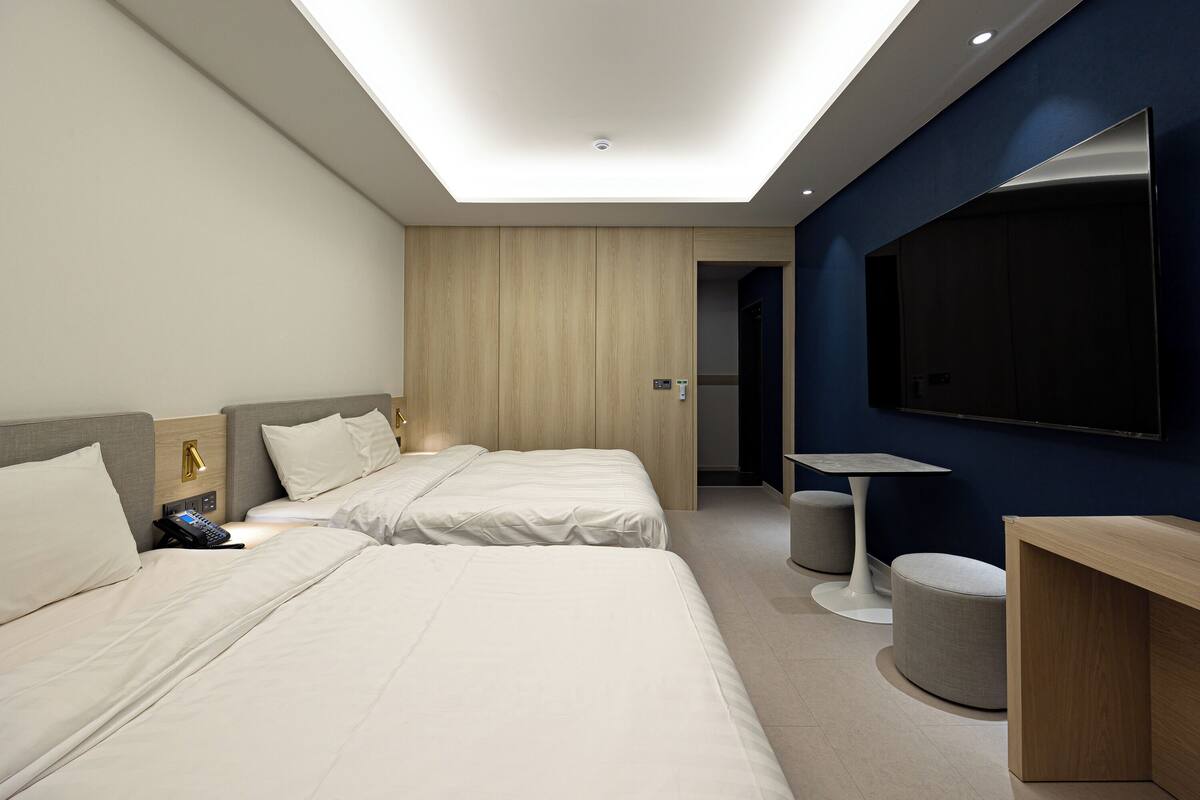 A modern hotel room with two beds, a desk, a chair, a TV, and a small seating area.