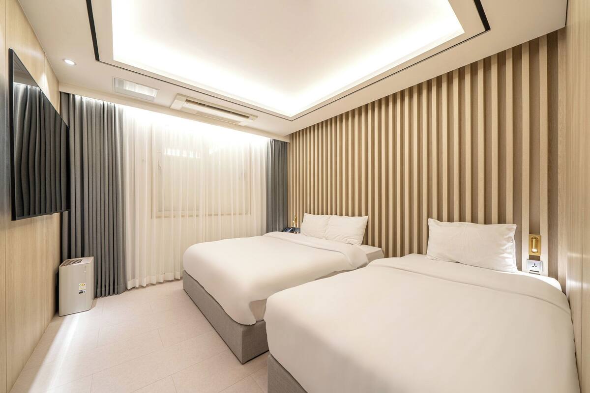 A modern hotel room with two beds, a flat-screen TV, a built-in air conditioner, and a large window with curtains.