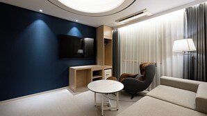 A modern hotel room with a dark blue accent wall, a flat-screen TV mounted above a wooden console, a massage chair, a sofa, and a round table. - Hound Hotel Gwangju Cheomdan (Gwangju)
