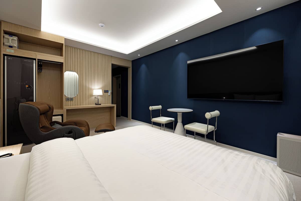 A modern hotel room with a large bed, a desk with a chair, a TV mounted on the wall, and a small table with two chairs.