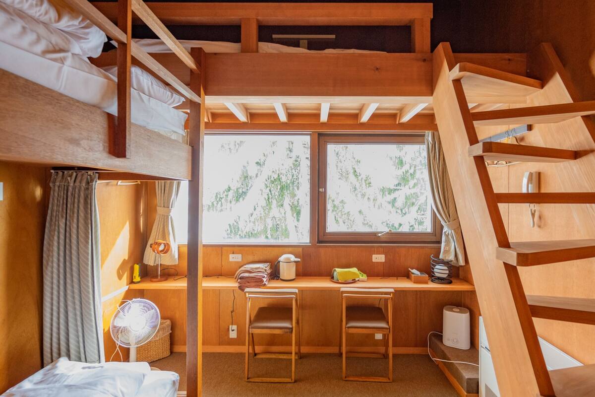 A compact room with a bunk bed, a desk with a chair, a fan, and a window with a view of greenery.