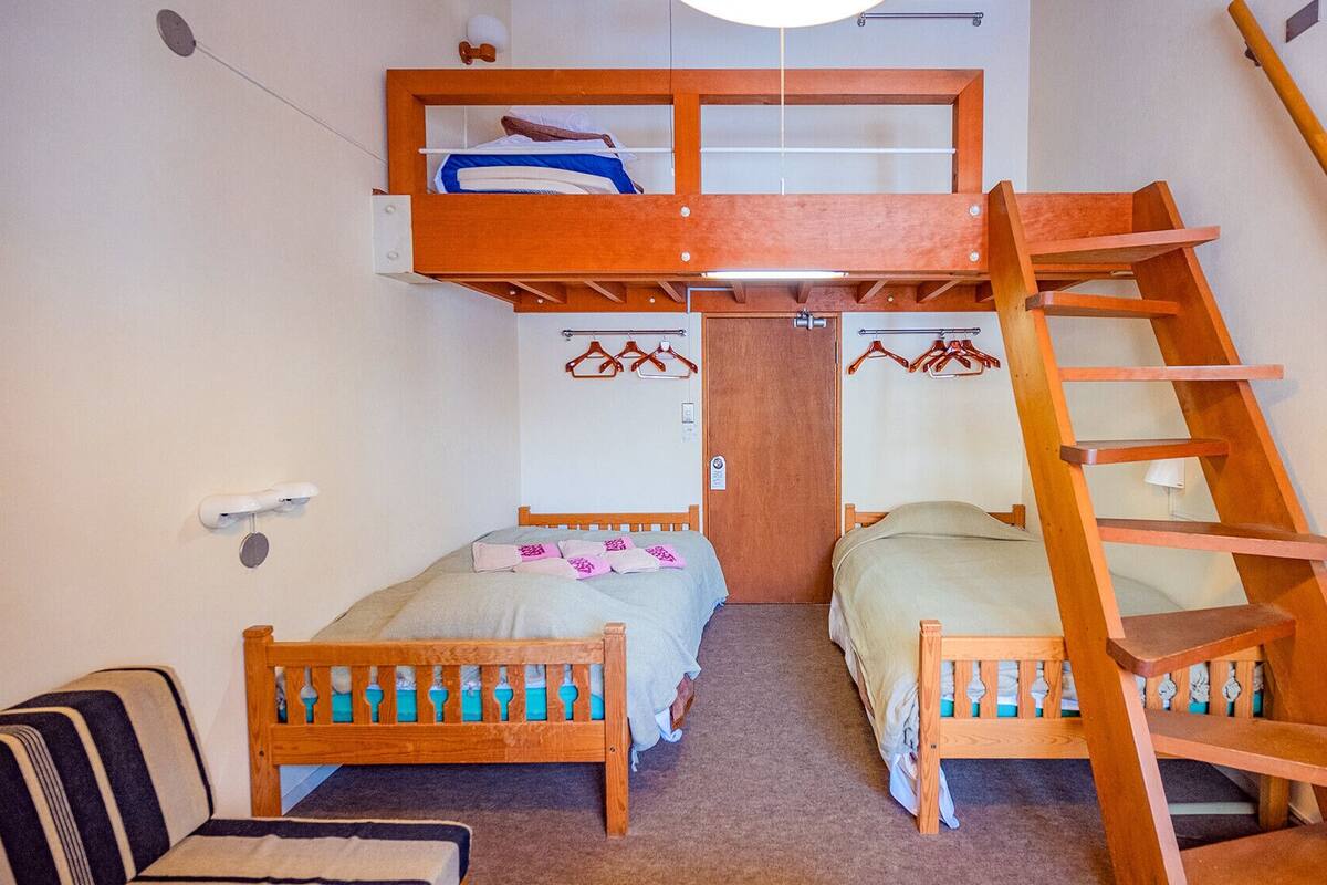 A dormitory room with two beds, a bunk bed, a wooden ladder, and a small seating area.