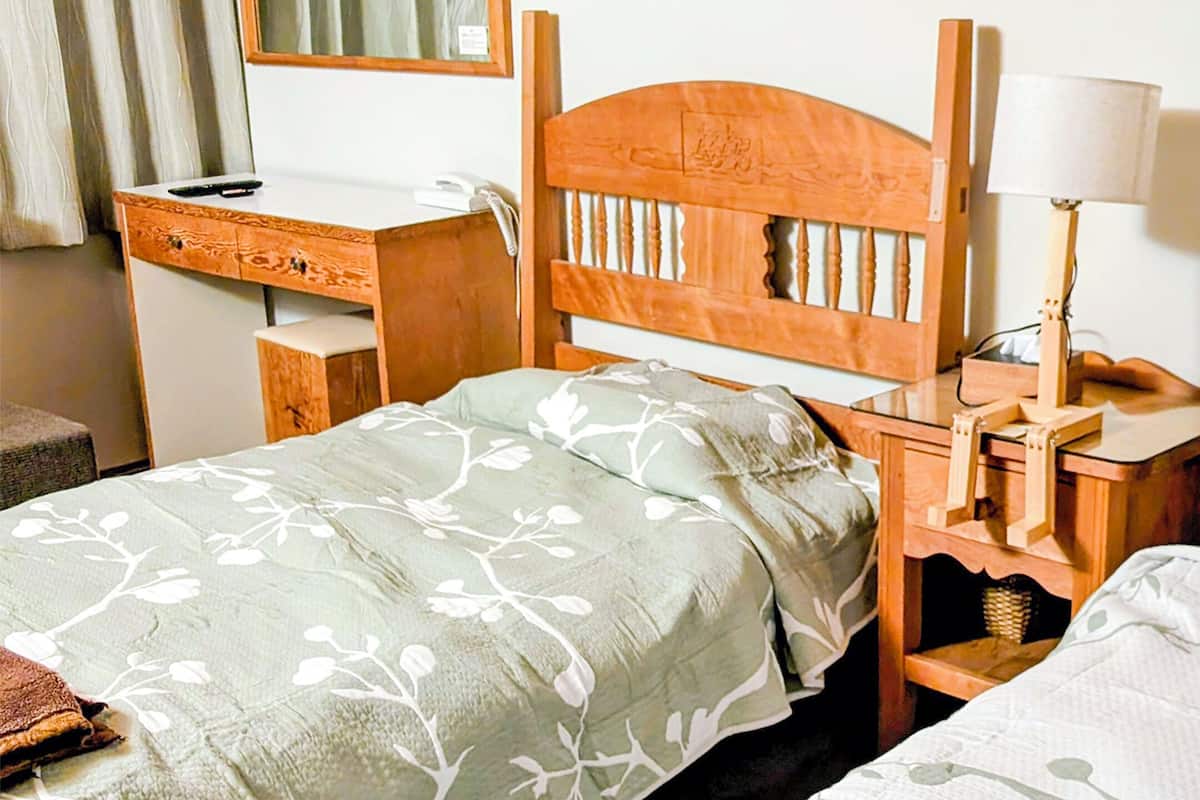 A hotel room with two beds, a wooden headboard, a bedside table with a lamp, a desk with a telephone, and a mirror on the wall.