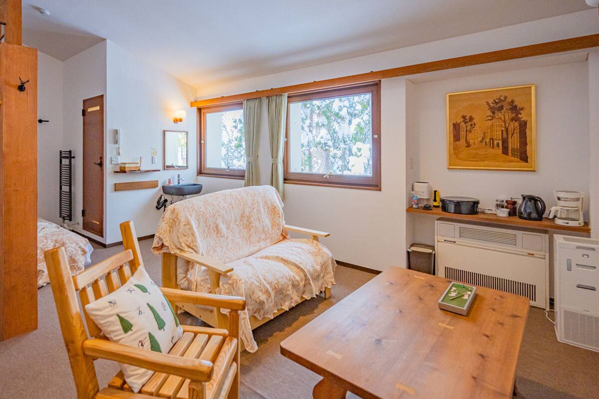A cozy room with bunk beds, a wooden table, a chair, a desk with a computer, and a window with curtains.