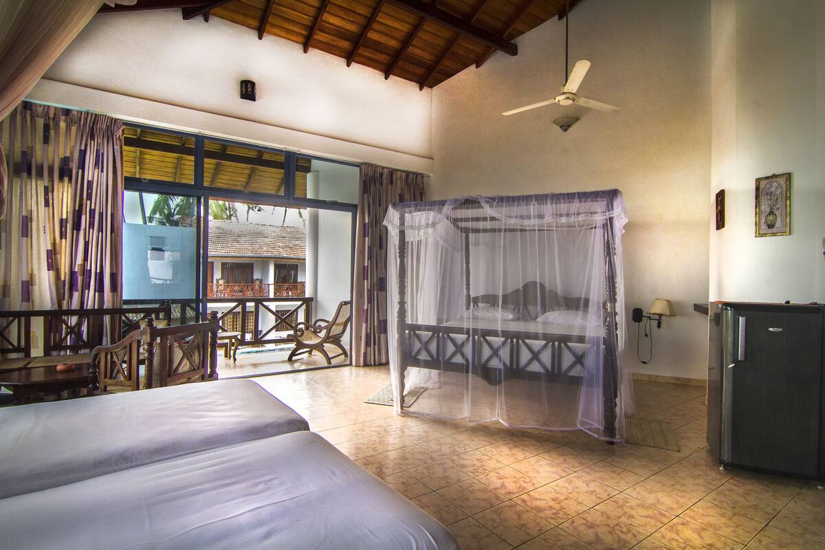 A bedroom with a bed, a bedside table, a chair, a mosquito net, a ceiling fan, and a window with curtains.