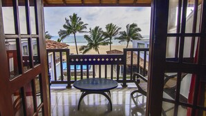 Un balcón abierto con una mesa y sillas, con vista a una playa con palmeras y un edificio al fondo.