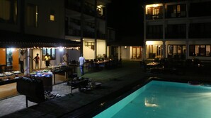 Zona de piscina exterior de noche con barbacoa y mesas de comedor.