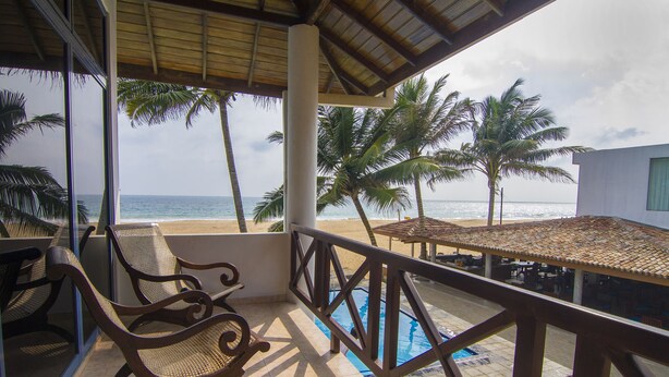 Un balcón con vista a la playa y al océano, una piscina y sillas de madera.