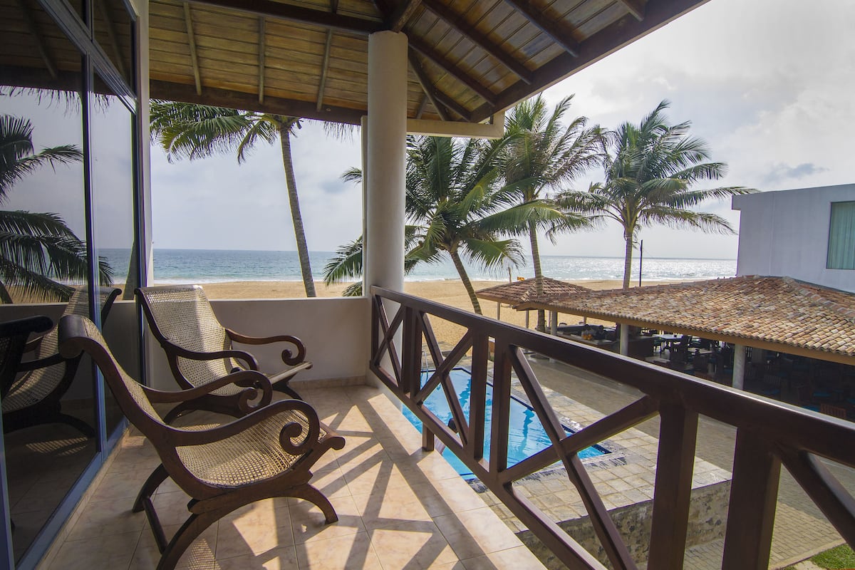A balcony with a view of the beach and ocean, a pool, and wooden chairs.