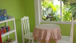 Quarto com parede verde, uma cadeira branca, uma mesa pequena com toalha xadrez e uma prateleira com bebidas e livros.