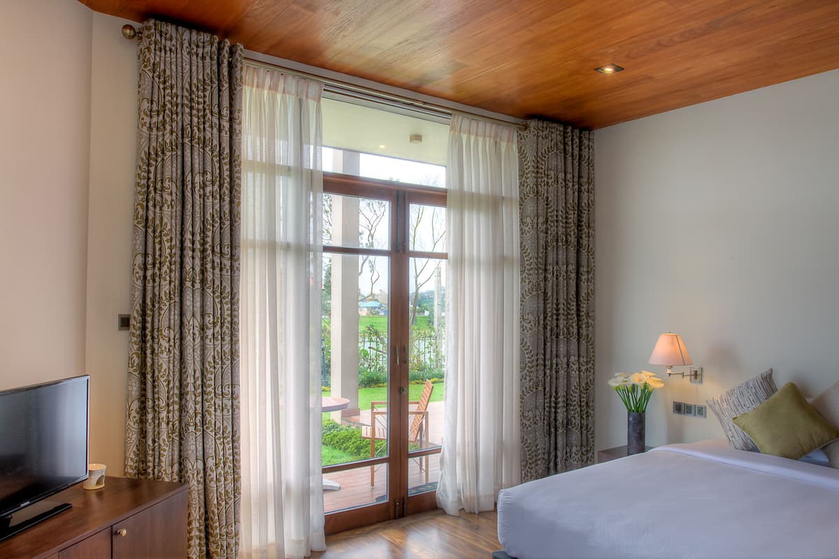 A bedroom with a bed, a TV on a wooden cabinet, a nightstand with a lamp, and a view of a garden through a glass door.