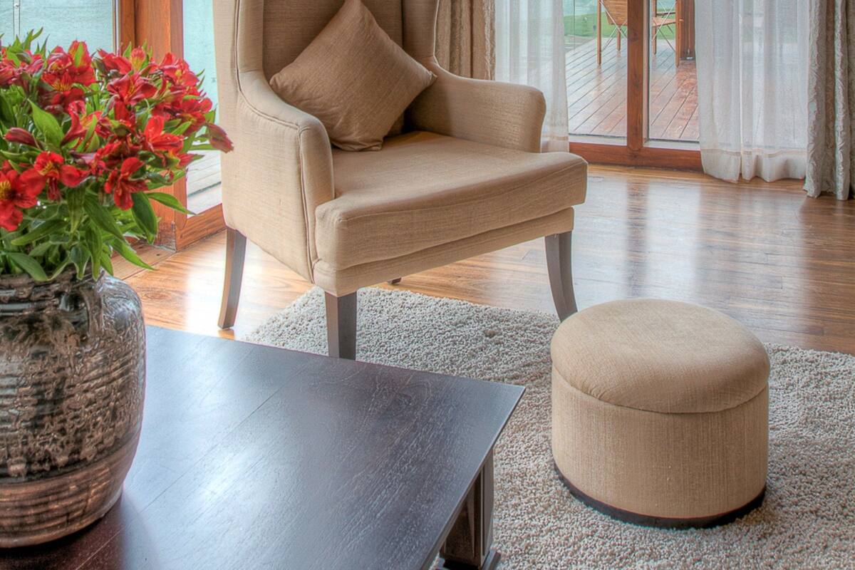 A room with a large window, a beige armchair, a small ottoman, a dark wooden table, and a vase with red flowers.
