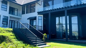 Una casa moderna de dos pisos con una escalera que lleva a un balcón, ventanas amplias y un césped cuidado.