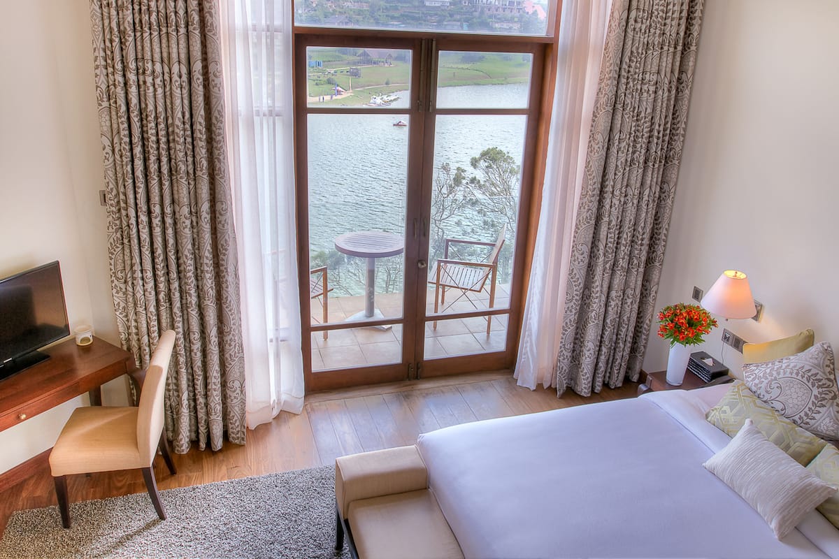 A hotel room with a bed, a desk with a TV, a chair, a table, and a view of a lake through a glass door.