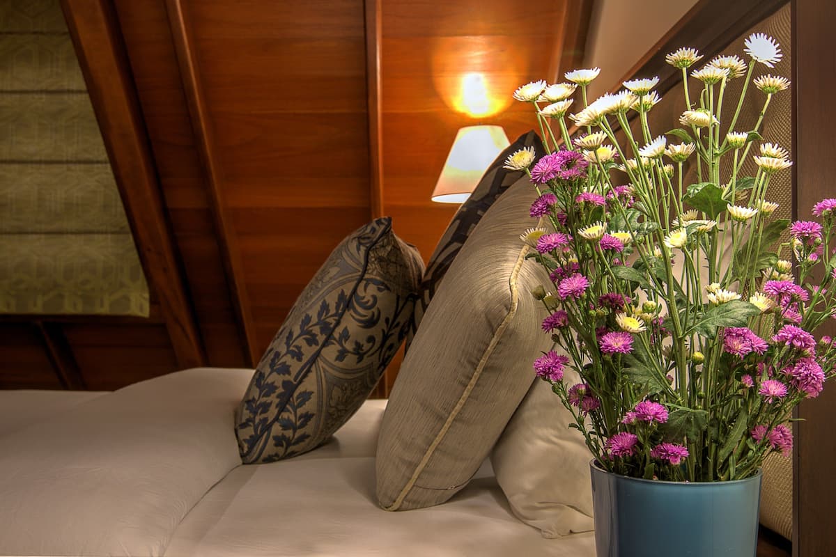 A bed with two pillows, a vase with flowers, and a lamp in the background.