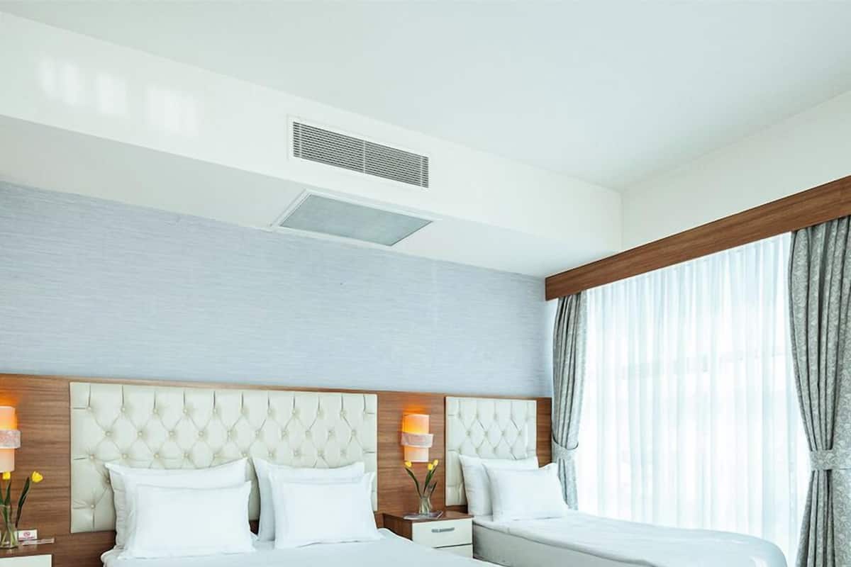 A hotel room with two beds, a wooden floor, a large window with curtains, and a ceiling vent.