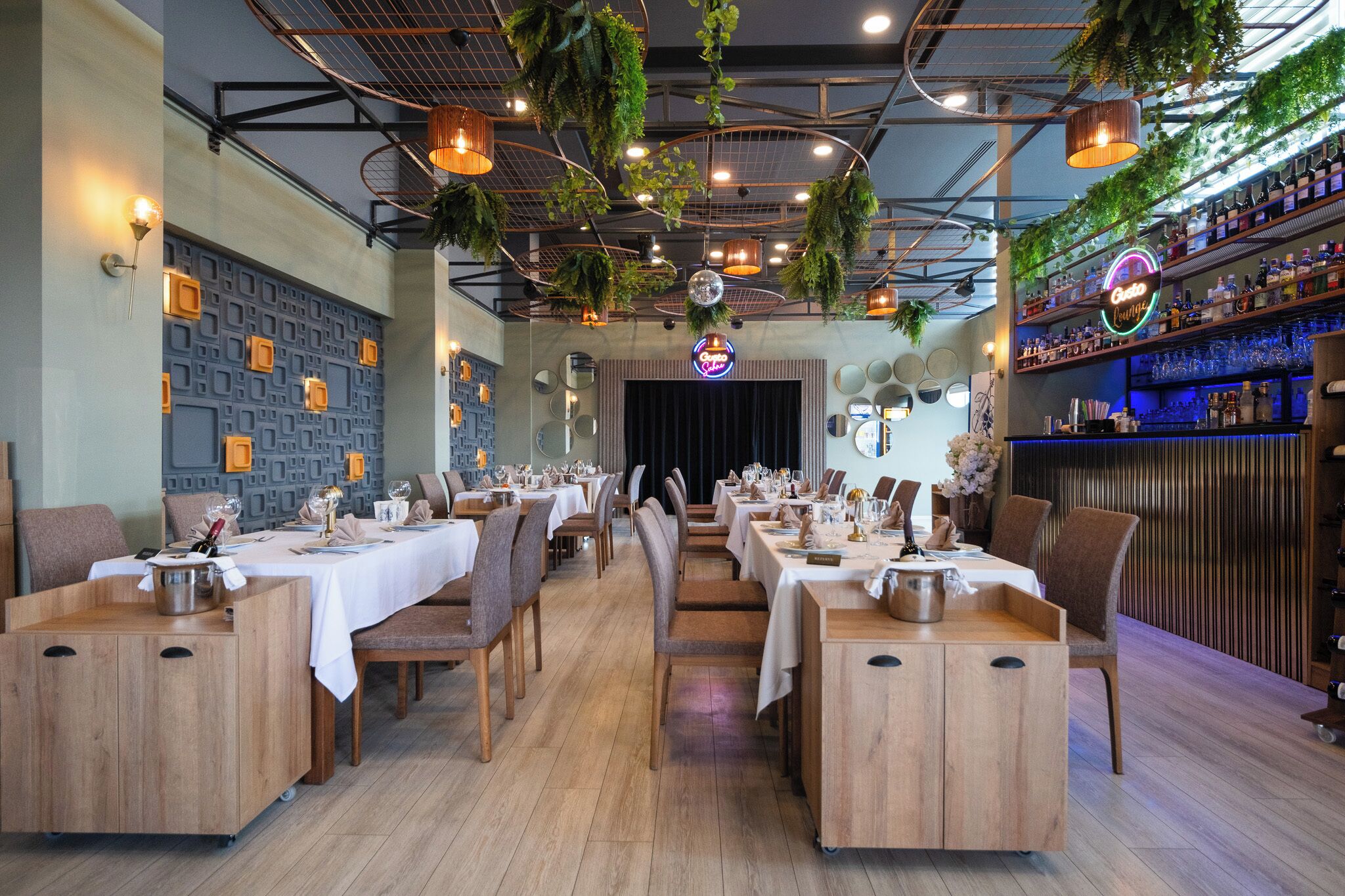 A restaurant with wooden tables and chairs, a bar area with shelves of bottles, and hanging plants.