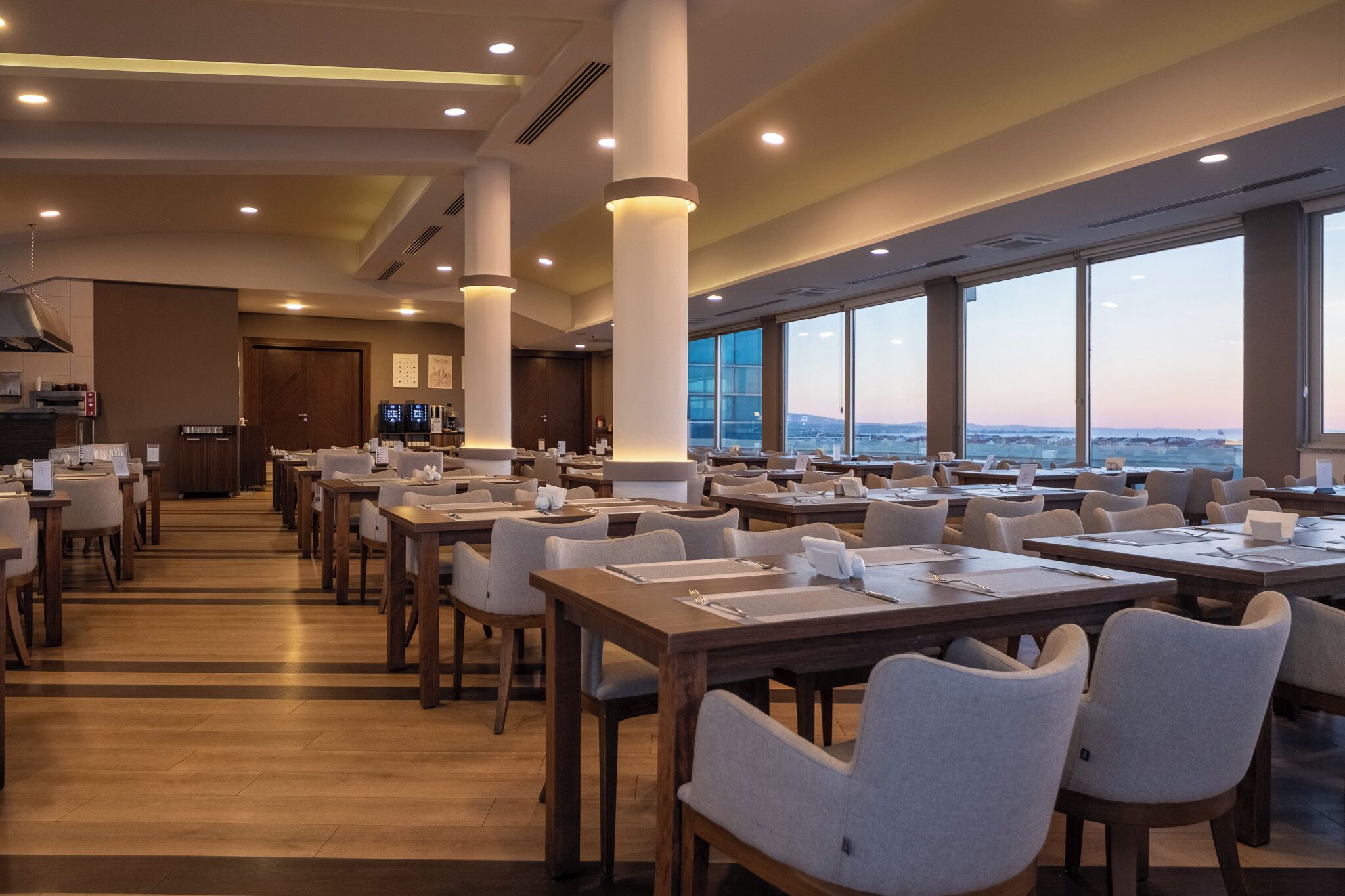 A spacious dining area with multiple tables and chairs, large windows offering a view of the cityscape, and modern lighting fixtures.