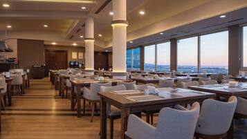 A spacious dining area with multiple tables and chairs, large windows offering a view of the cityscape, and modern lighting fixtures.