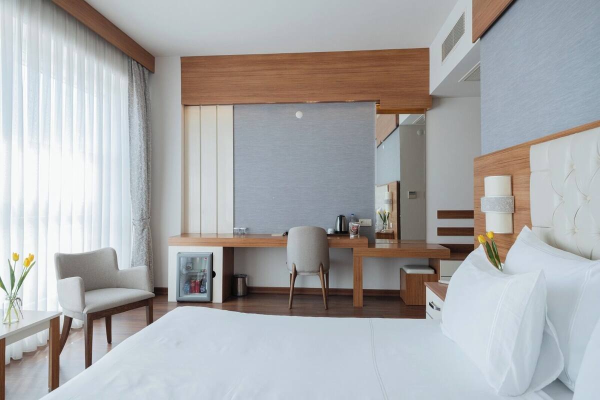A hotel room with a bed, a desk, a chair, a small fridge, and a bedside table with a lamp.