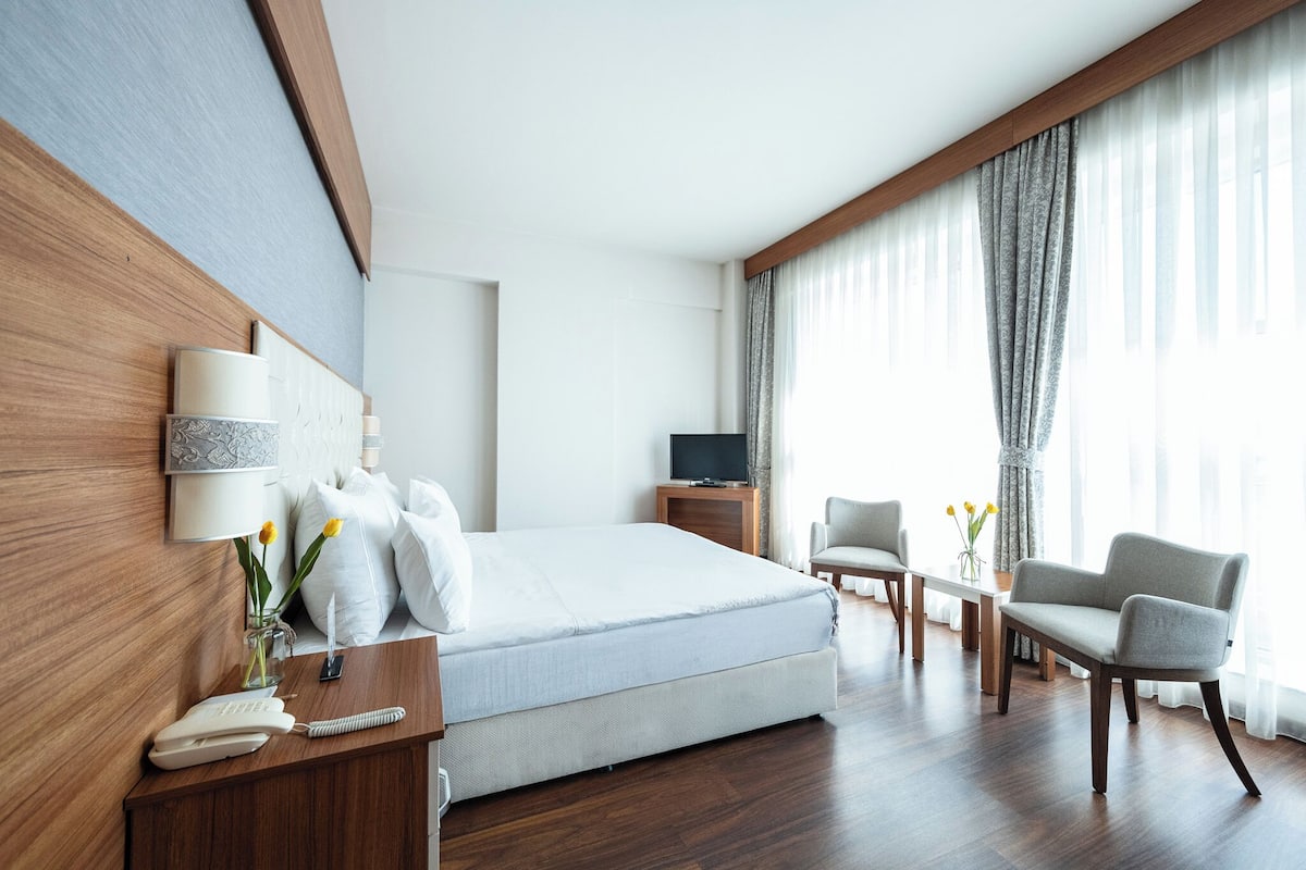 A hotel room with a bed, bedside table, lamp, chair, TV, and a view of the outdoors through large windows.