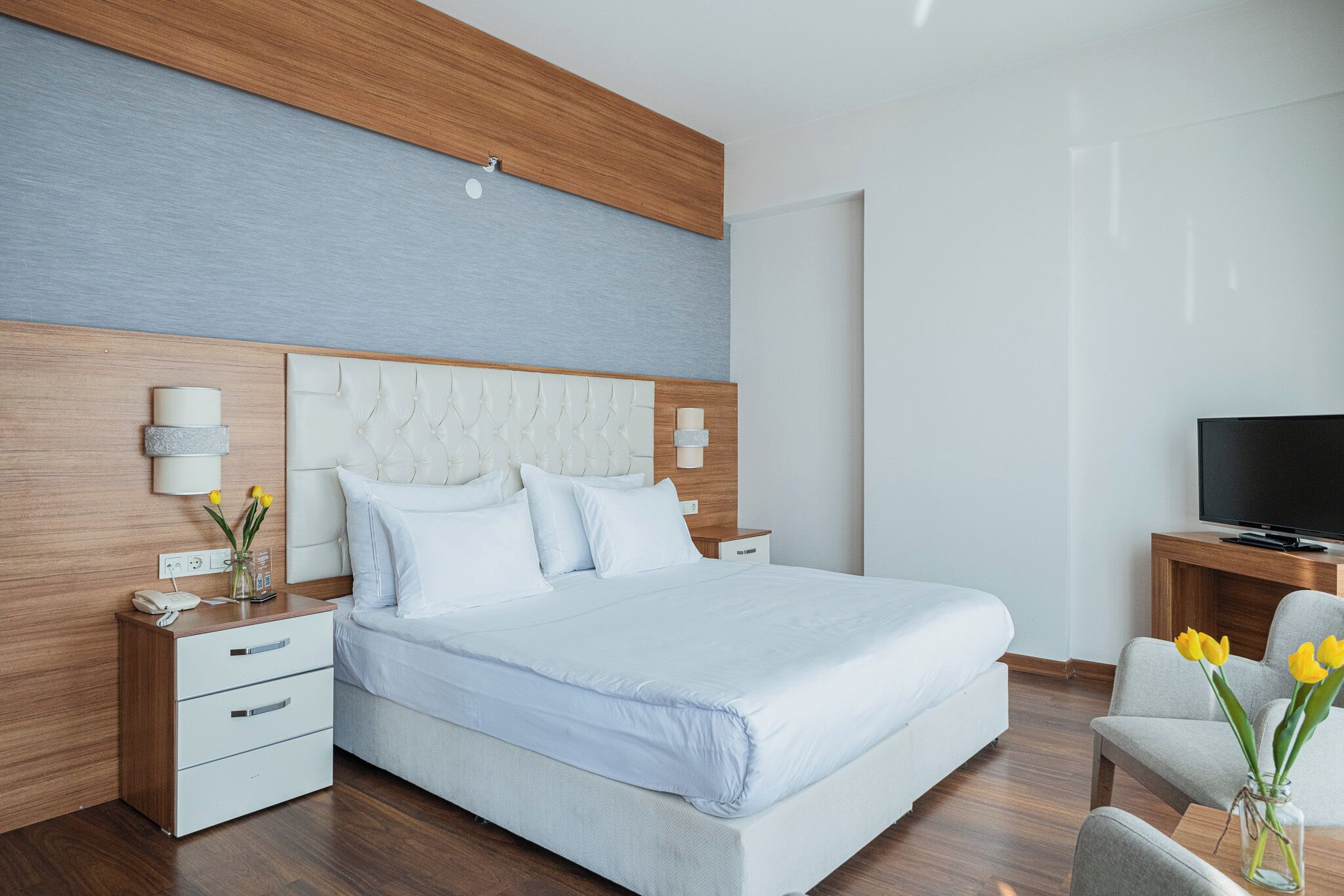 Una habitación de hotel moderna con una cama grande, cabecero de madera, mesitas de noche con lámparas, un televisor y un jarrón con flores.