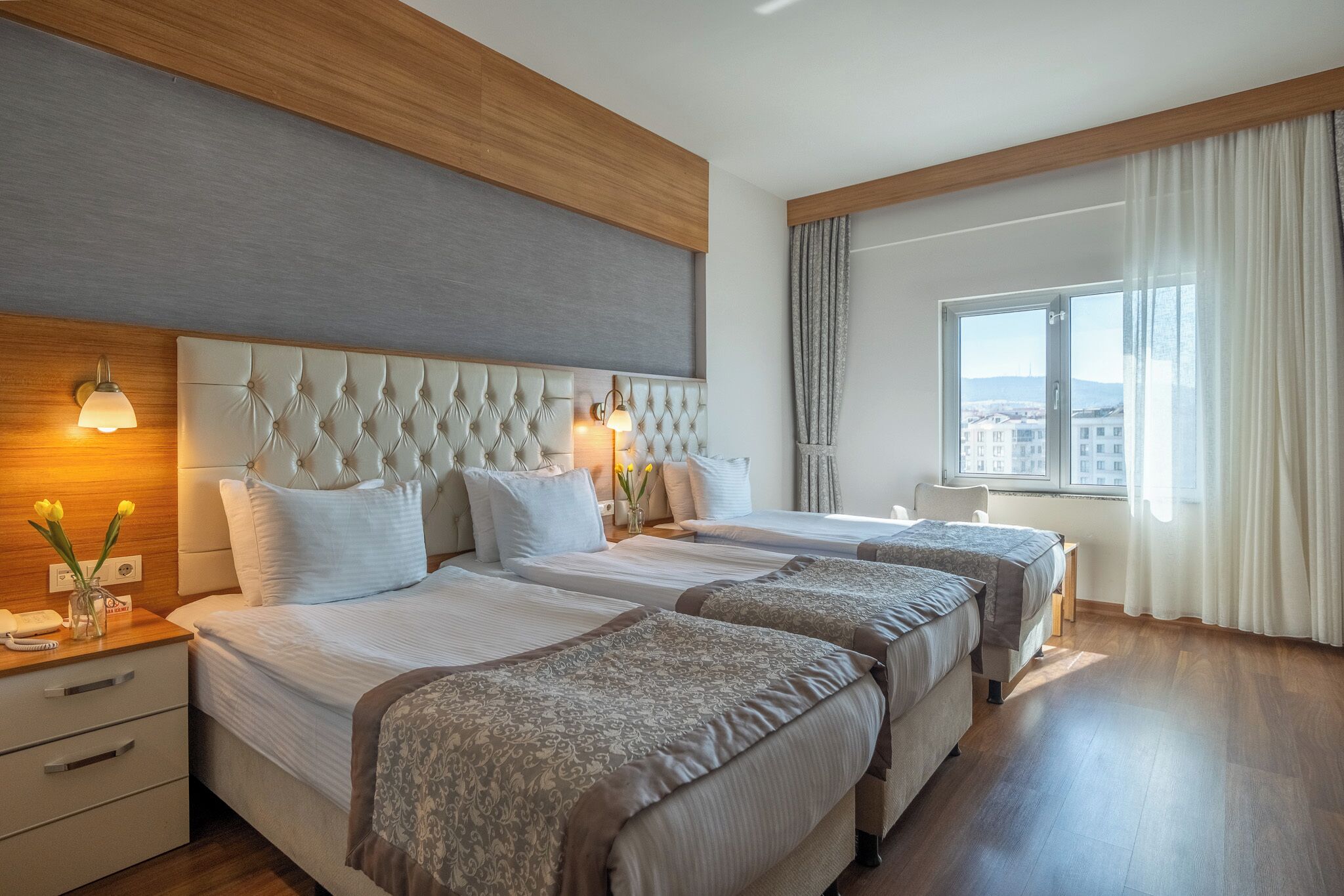 Habitación de hotel con dos camas, cabecero de madera, lámparas de noche, una ventana con cortinas y vistas a edificios.