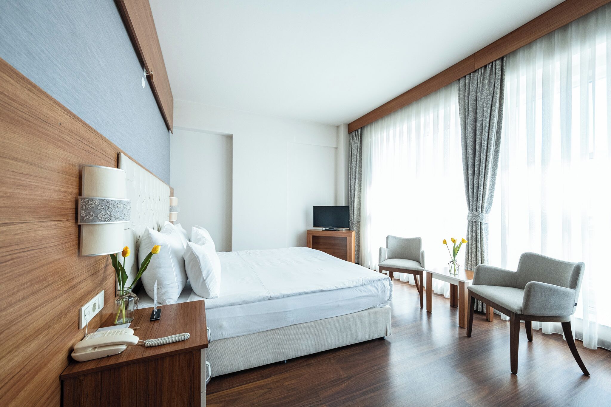 Ein Hotelzimmer mit Bett, Nachttisch, Fernseher, Sessel und Schreibtisch mit Telefon und Blumenvase.
