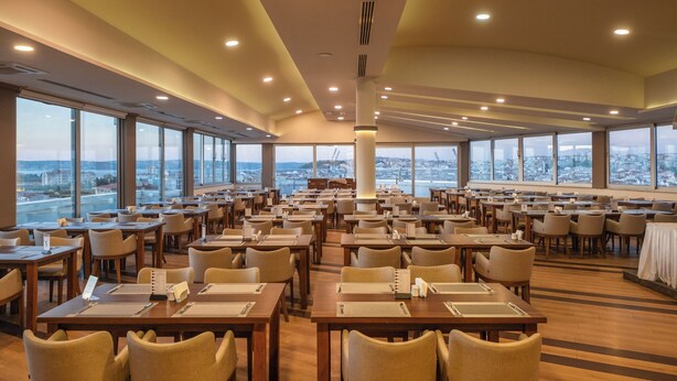 A spacious dining area with multiple tables and chairs, large windows offering a city view, and a central pillar.
