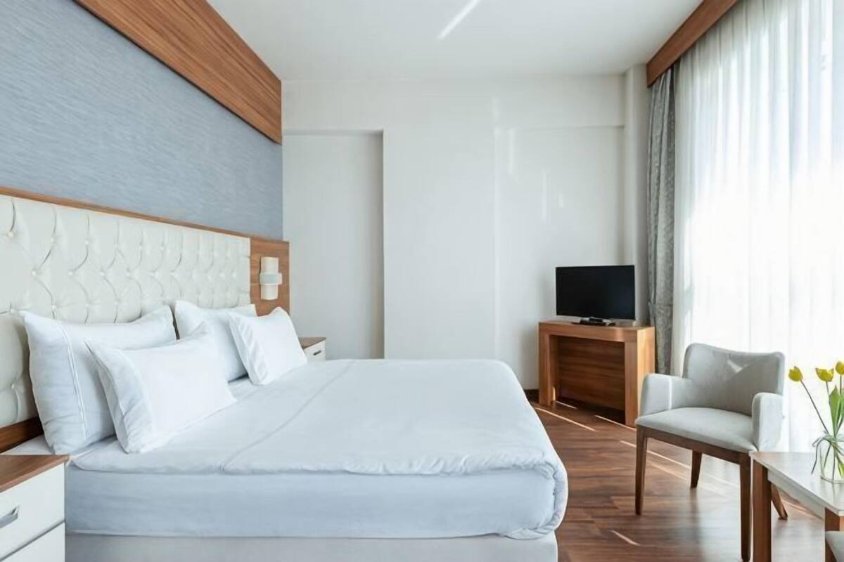 A hotel room with a large bed, a wooden headboard, a television on a wooden stand, a chair, a small table with a vase, and a window with curtains.
