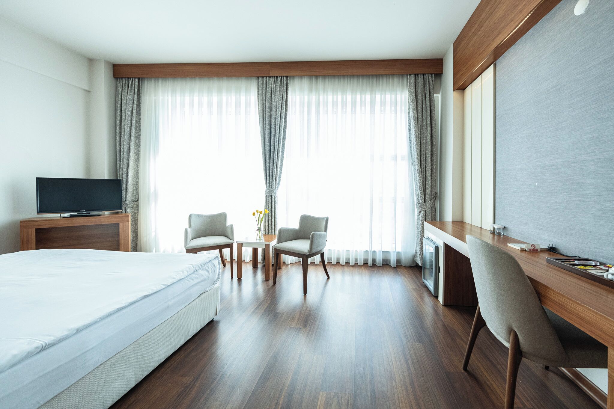 A hotel room with a bed, a desk, two chairs, a TV, and large windows with curtains.
