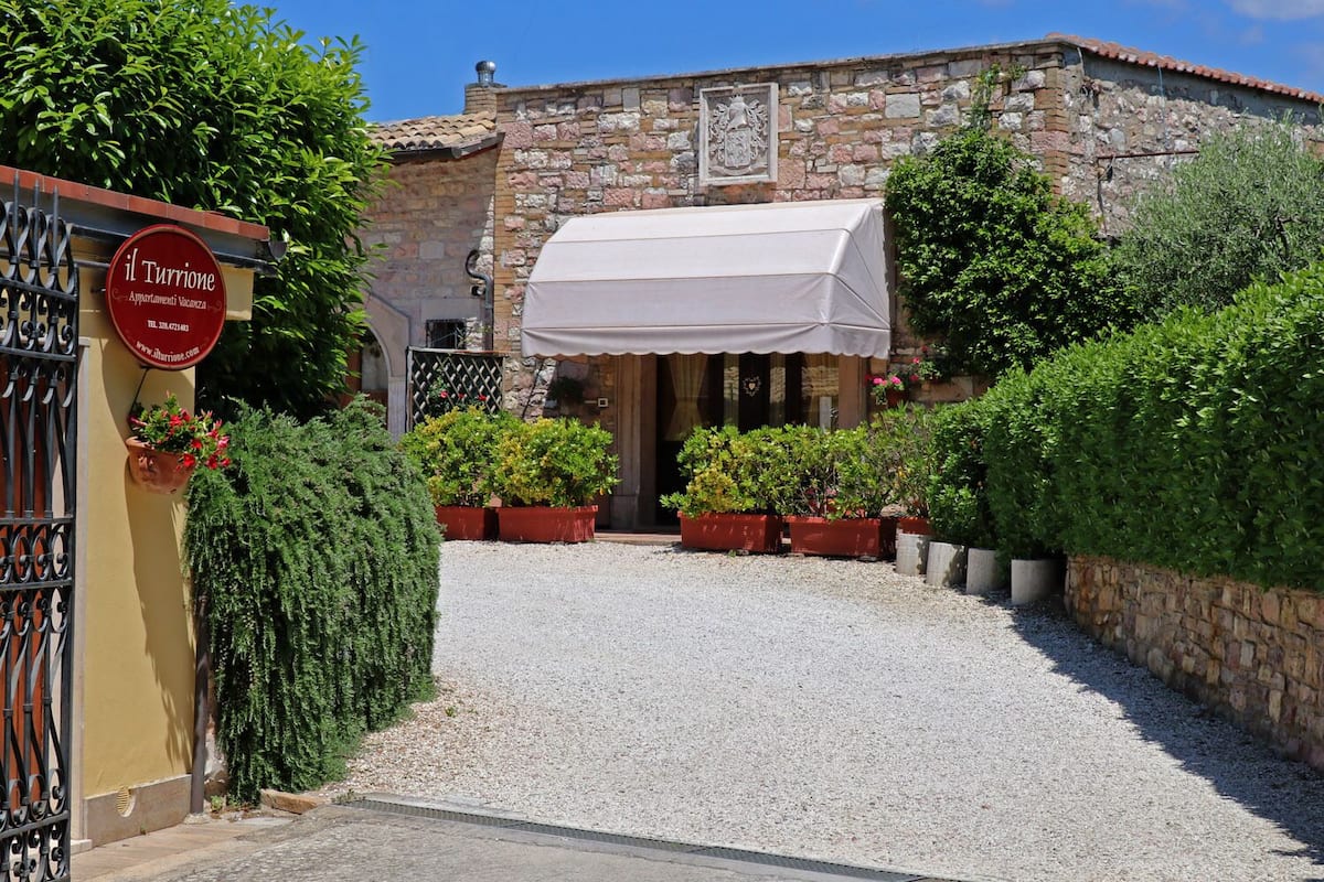 Un bâtiment en pierre avec une allée de gravier menant à une entrée abritée. On y trouve des plantes en pot et un panneau indiquant qu’il s’agit d’une location de vacances.