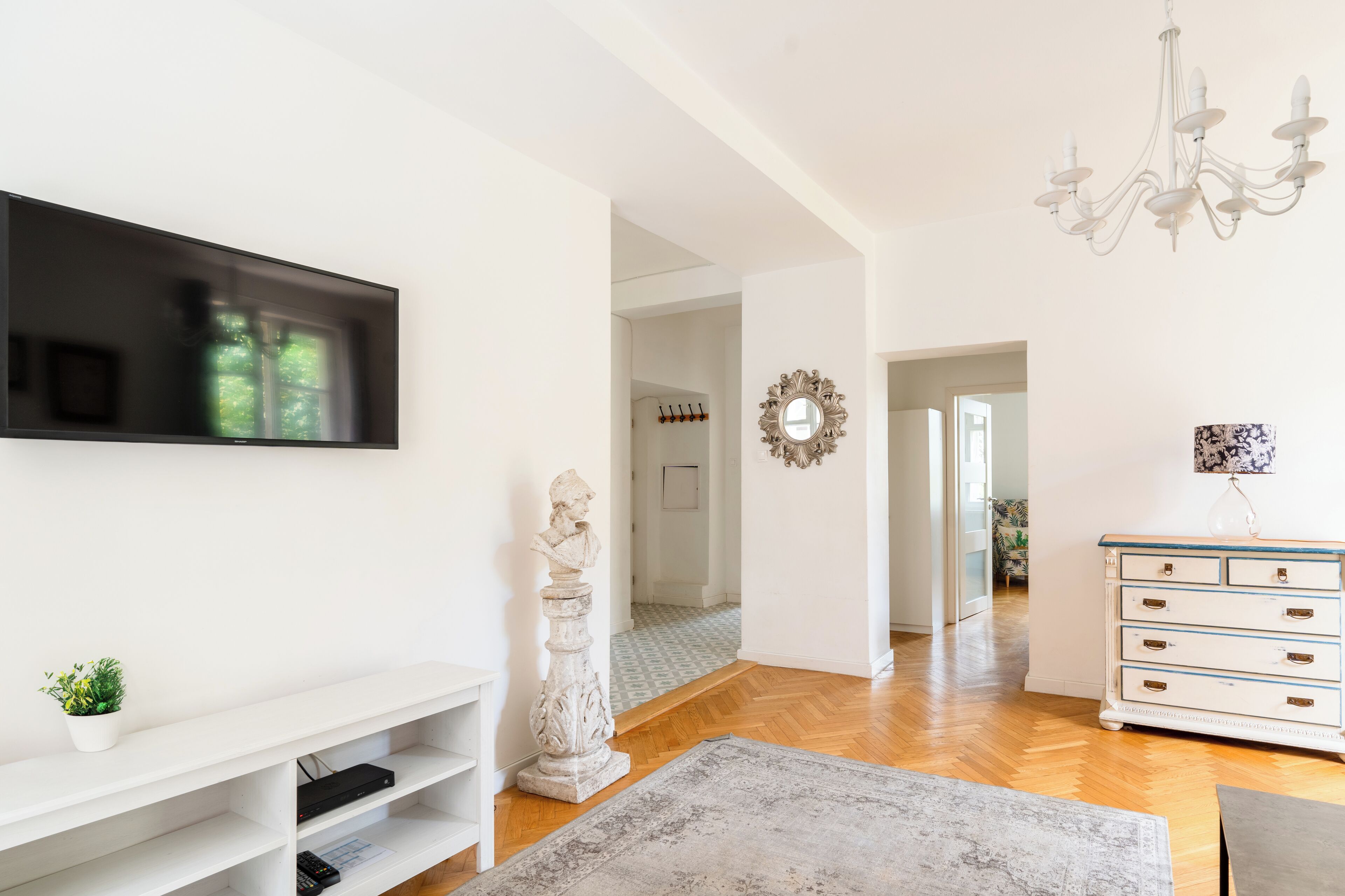 Una sala moderna con un televisor de pantalla plana, una consola blanca, una estatua, un candelabro y un pasillo que lleva a otra habitación.